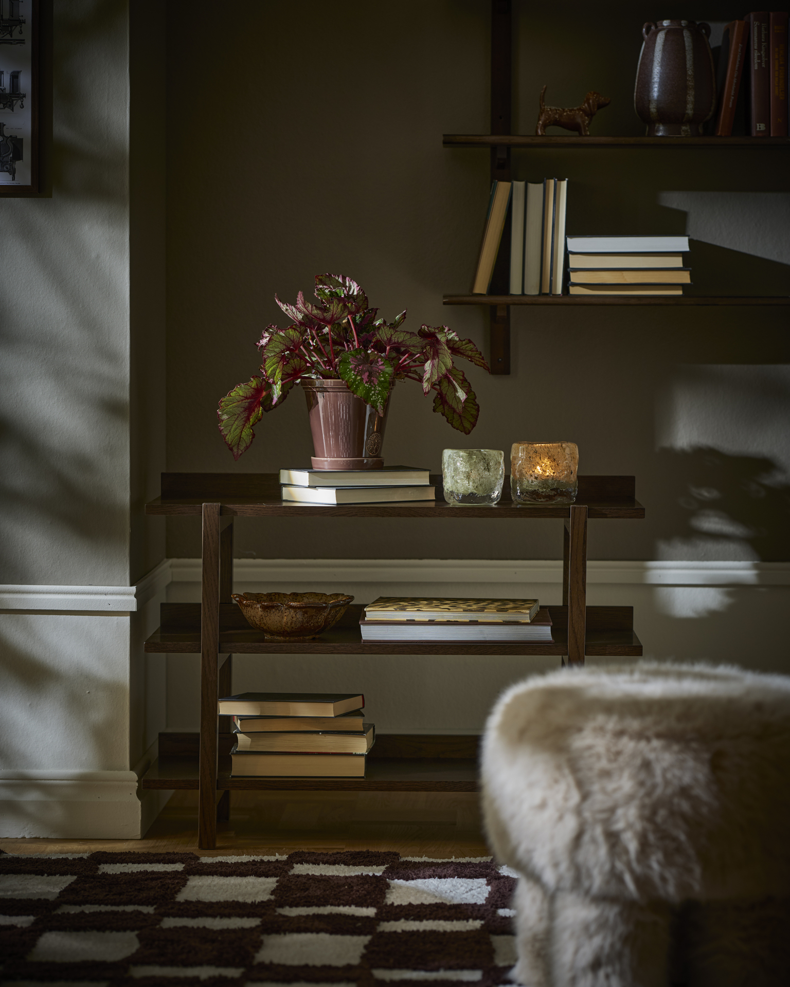En hylla av mörkbrun ekfanér med tre hyllplan, står i ett rum med mörka toner. På hyllorna står en blomma, två ljuslyktor, en skål och böcker. På bilden syns även en vägghylla och en grå sittpuff.