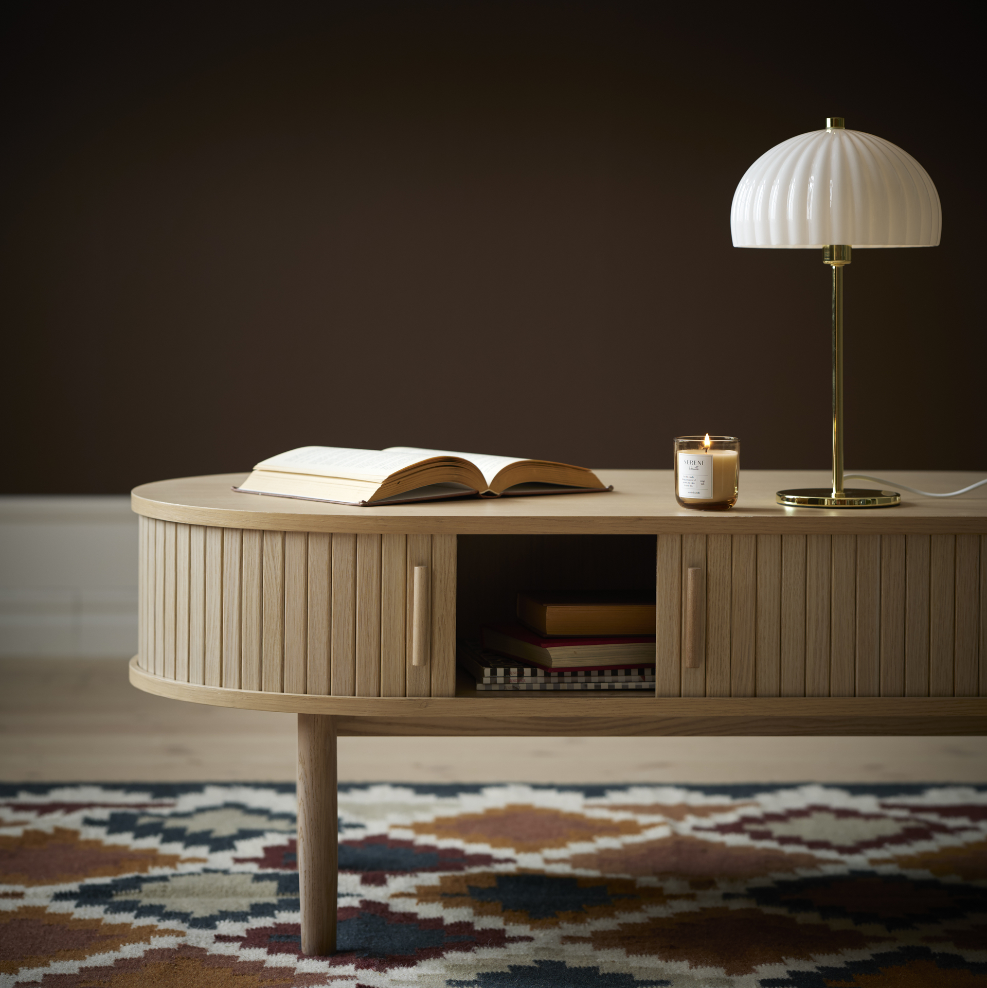 Ett ovalt, ribbat oval soffbord av ekfanér står på en rutig matta. På bordet finns en bok, en lampa och ett tänt doftljus. I bakgrunden syns en brun tapet.