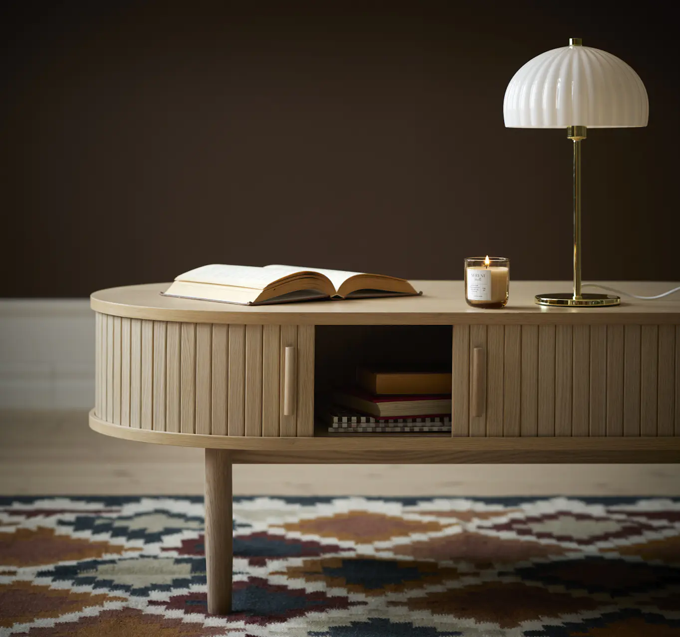 Ett ovalt, ribbat oval soffbord av ekfanér står på en rutig matta. På bordet finns en bok, en lampa och ett tänt doftljus. I bakgrunden syns en brun tapet.