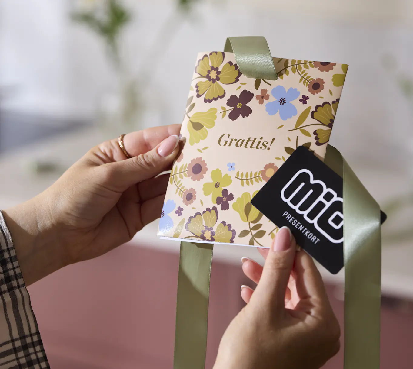 Två händer håller i ett presentkort från Mio och ett konvolut med ett blommigt mönster i naturnära toner