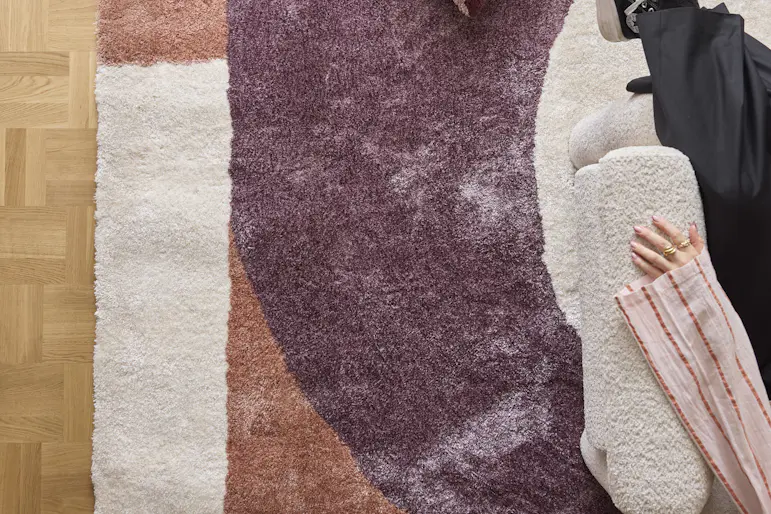 En vit, lila och rosa matta med asymmetriskt mönster. På bilden syns även en randig kudde och ett par ben.