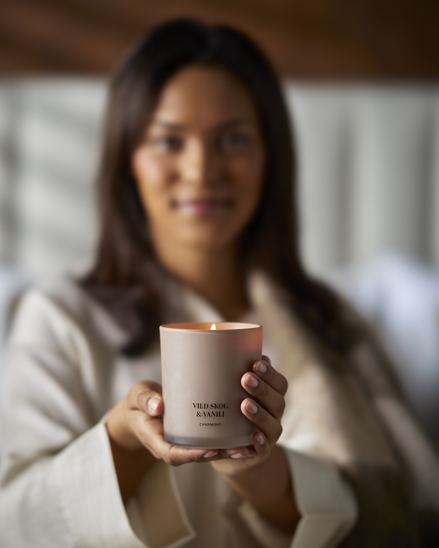 En brunhårig tjej sitter i en säng och håller fram ett rosa doftljus 