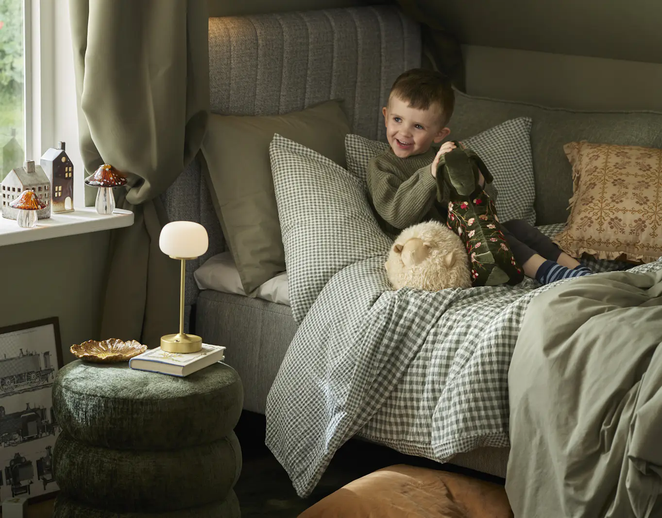 I en säng bäddad med gröna sängkläder i olika mönster sitter en pojke och håller i gosedjur föreställande ett får och en tax.
