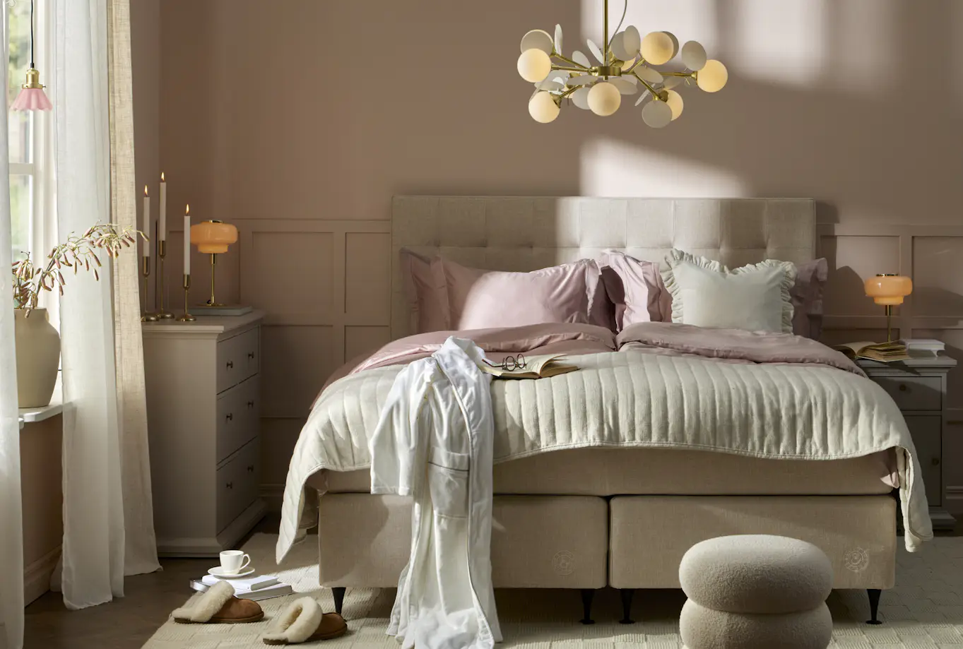En beige säng med rosa och vita sängkläder står i ett sovrum med ljusrosa väggar.