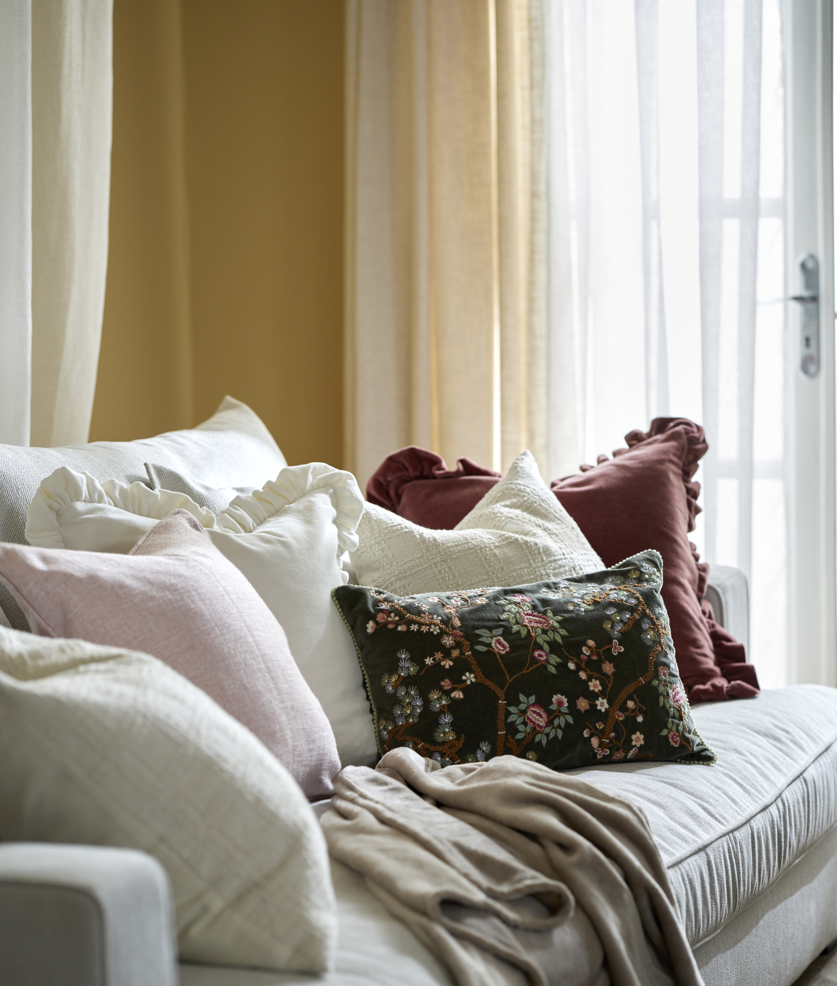På en ljus soffa ligger flera vita kuddar, en mörkröd kudde med volang, en ljusrosa kudde och en mörkgrön kudde med borderi. I bakgrunden hänger en gul och en vit gardin.