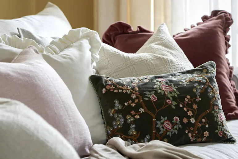 På en ljus soffa ligger flera vita kuddar, en mörkröd kudde med volang, en ljusrosa kudde och en mörkgrön kudde med borderi. I bakgrunden hänger en gul och en vit gardin.