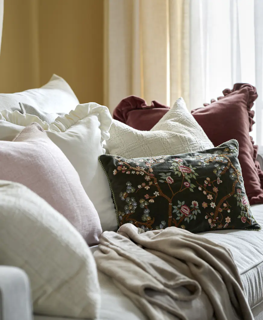 På en ljus soffa ligger flera vita kuddar, en mörkröd kudde med volang, en ljusrosa kudde och en mörkgrön kudde med borderi. I bakgrunden hänger en gul och en vit gardin.