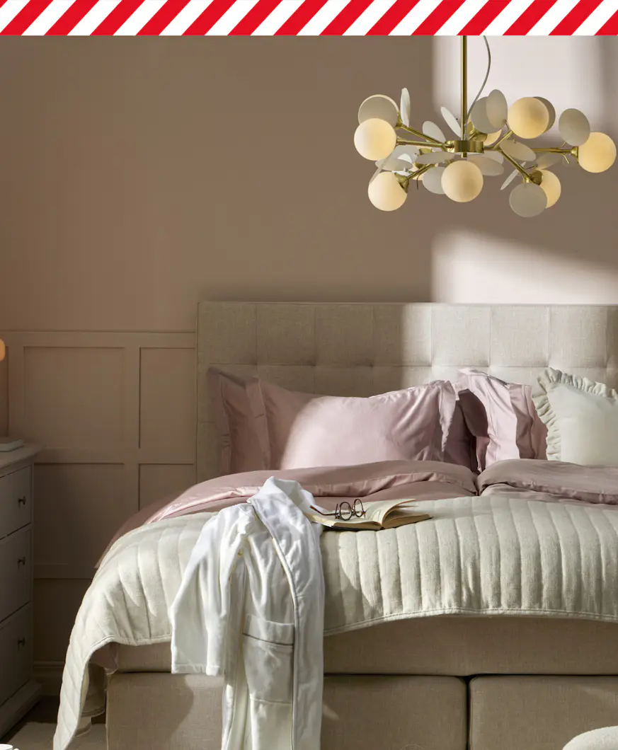 En beige säng med rosa och vita sängkläder står i ett sovrum med ljusrosa väggar. I bildens överkant finns en rödvit tejpremsa.