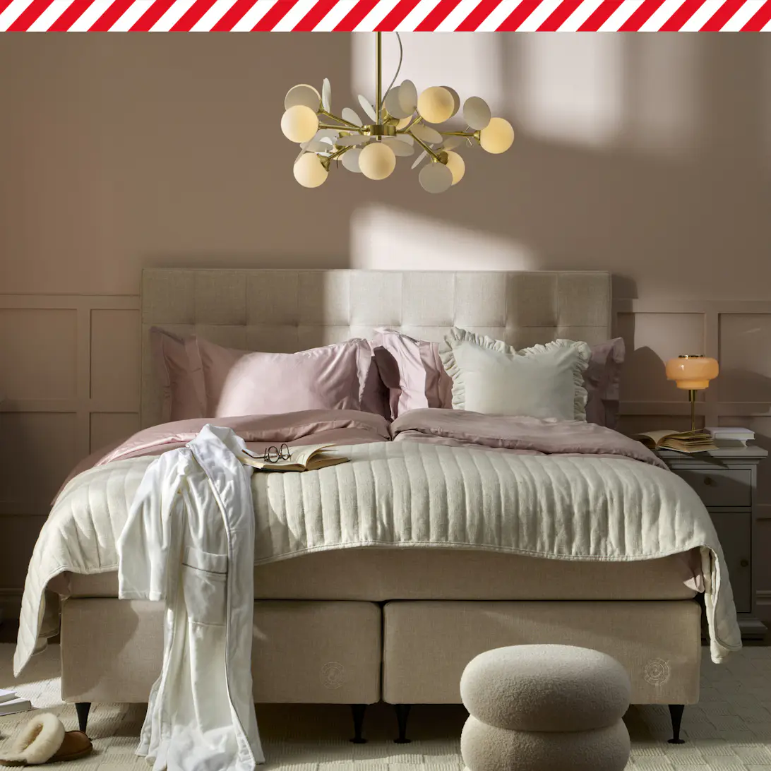 En beige säng med rosa och vita sängkläder står i ett sovrum med ljusrosa väggar. I bildens överkant finns en rödvit tejpremsa.