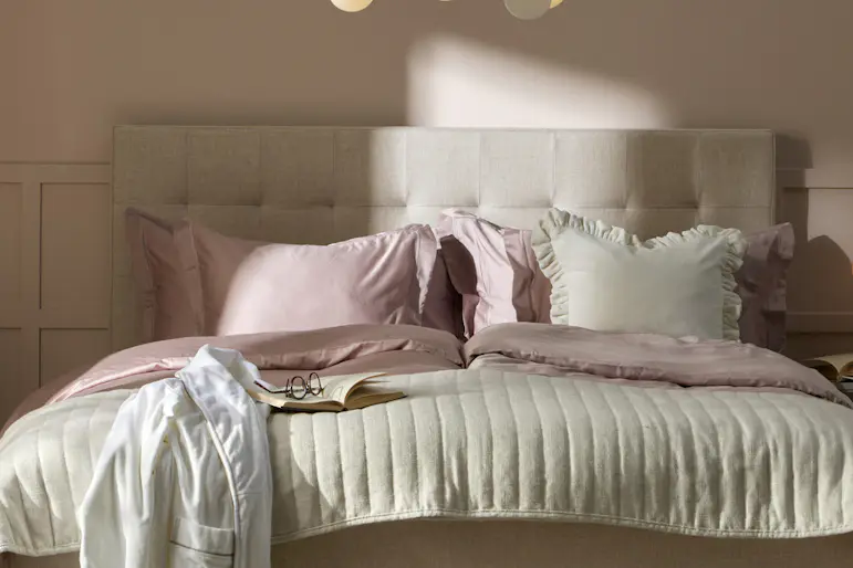 En beige säng med rosa och vita sängkläder står i ett sovrum med ljusrosa väggar. I bildens överkant finns en rödvit tejpremsa.