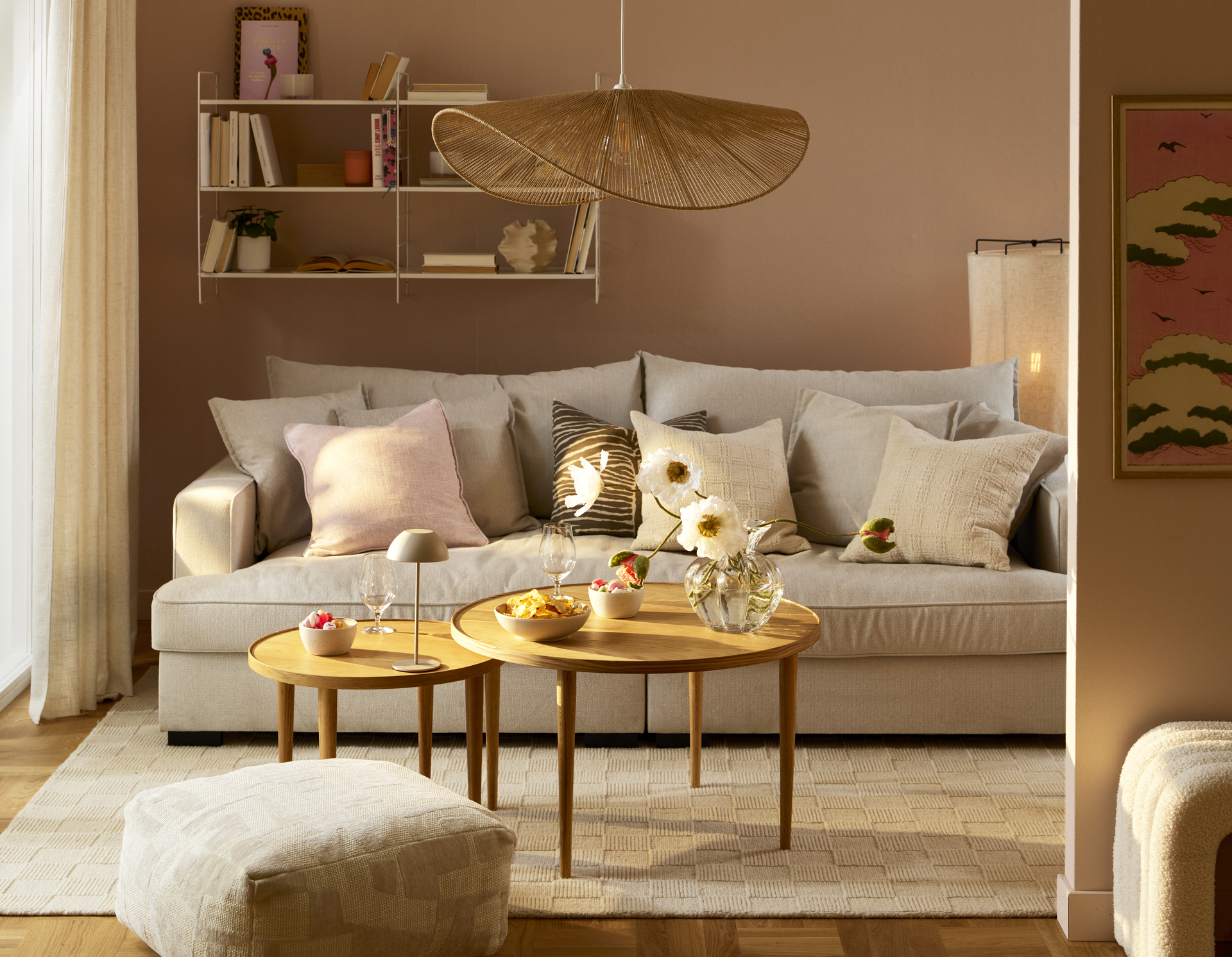 Beige soffa med flera vita, rosa och mönstrade kuddar står bakom ett satsbord i ett vardagsrum med ljusrosa väggar.