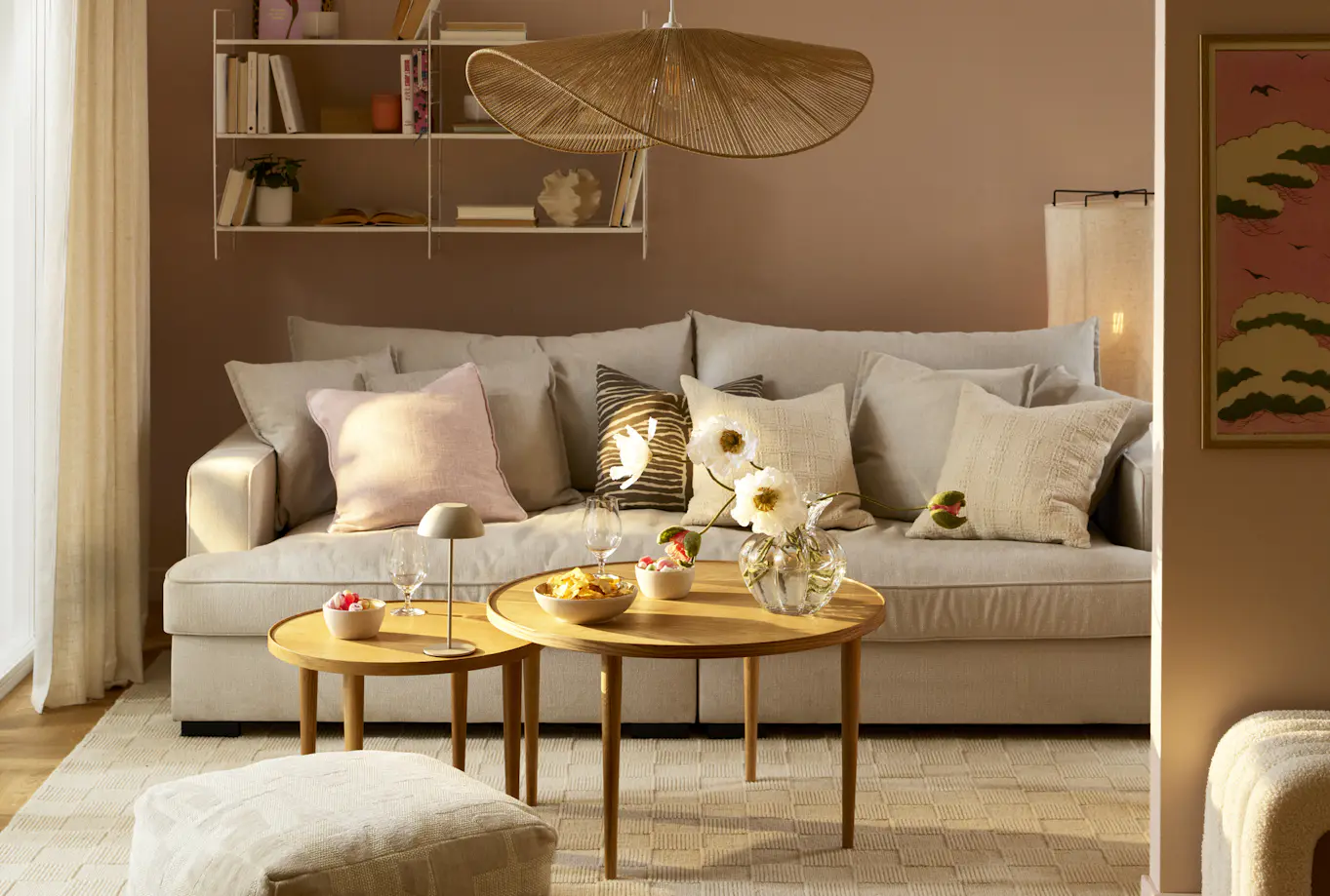 Beige soffa med flera vita, rosa och mönstrade kuddar står bakom ett satsbord i ett vardagsrum med ljusrosa väggar.