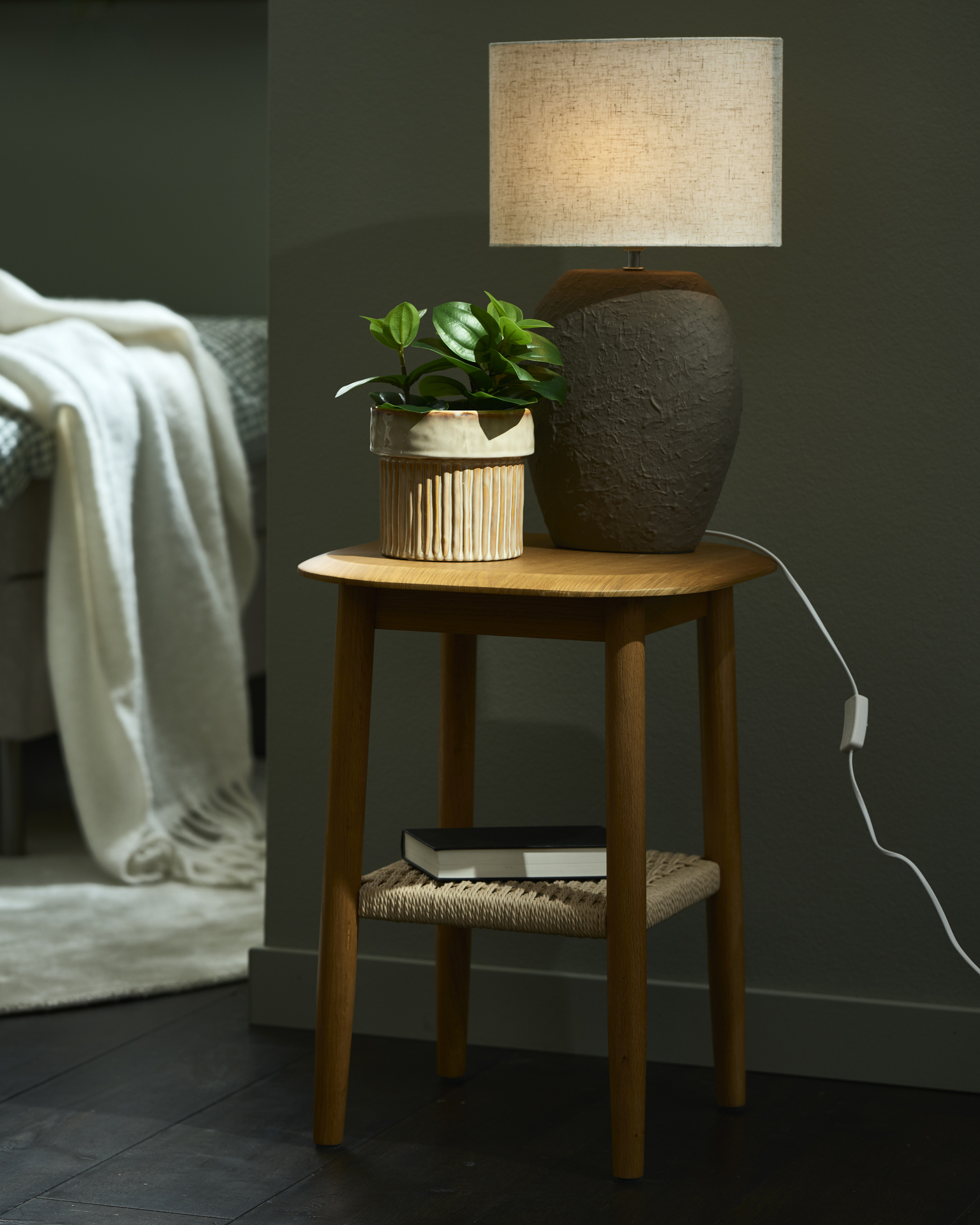 Ett sidobord av lackerad ek med en flätad underhylla. På bordet står en bordslampa och en krukväxt.
