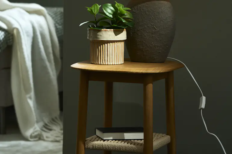 Ett sidobord av lackerad ek med en flätad underhylla. På bordet står en bordslampa och en krukväxt.