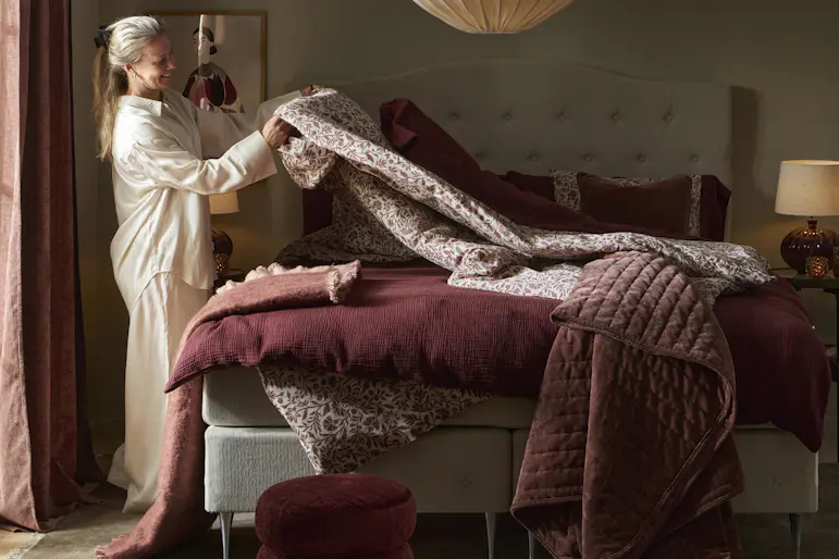 En tjej i pyjamas står i ett sovrum och bäddar en säng med röda påslakan
