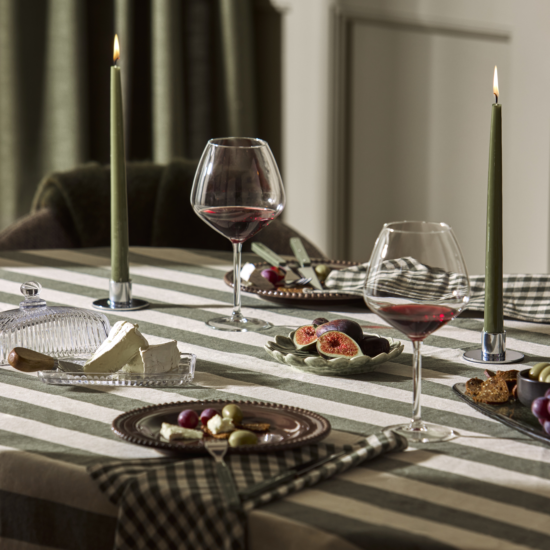 Ett uppdukat bord med en grön-/vitrandig duk, brun assiett, rutiga servetter, vinglas och stearinljus.