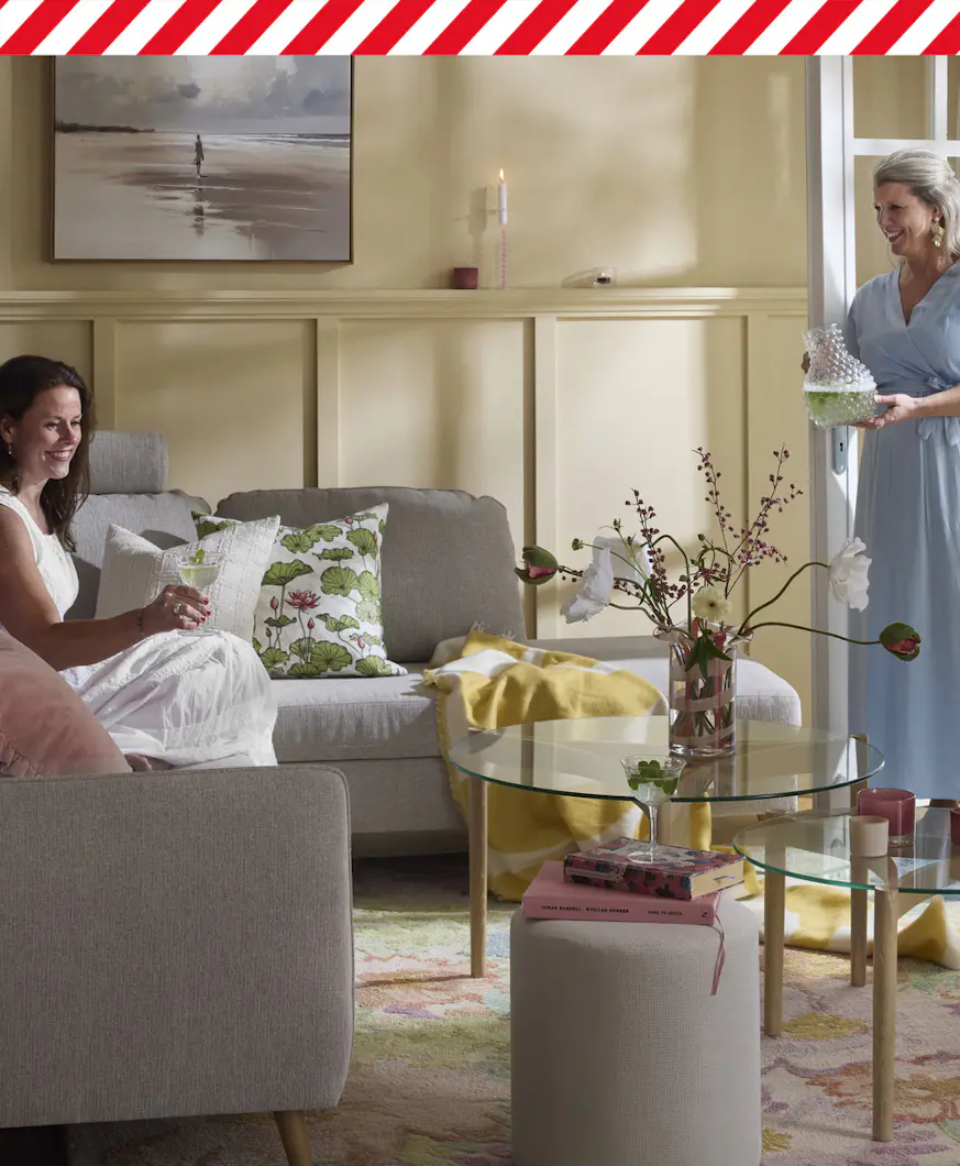 Kvinna sitter i grå 2,5-sitssoffa med divan som står på en blommig matta i ett vardagsrum med ljusgula väggar. Vid en altandörr står en annan kvinna och håller i en karaff. I bildens överkant går en röd och vit tejpremsa.