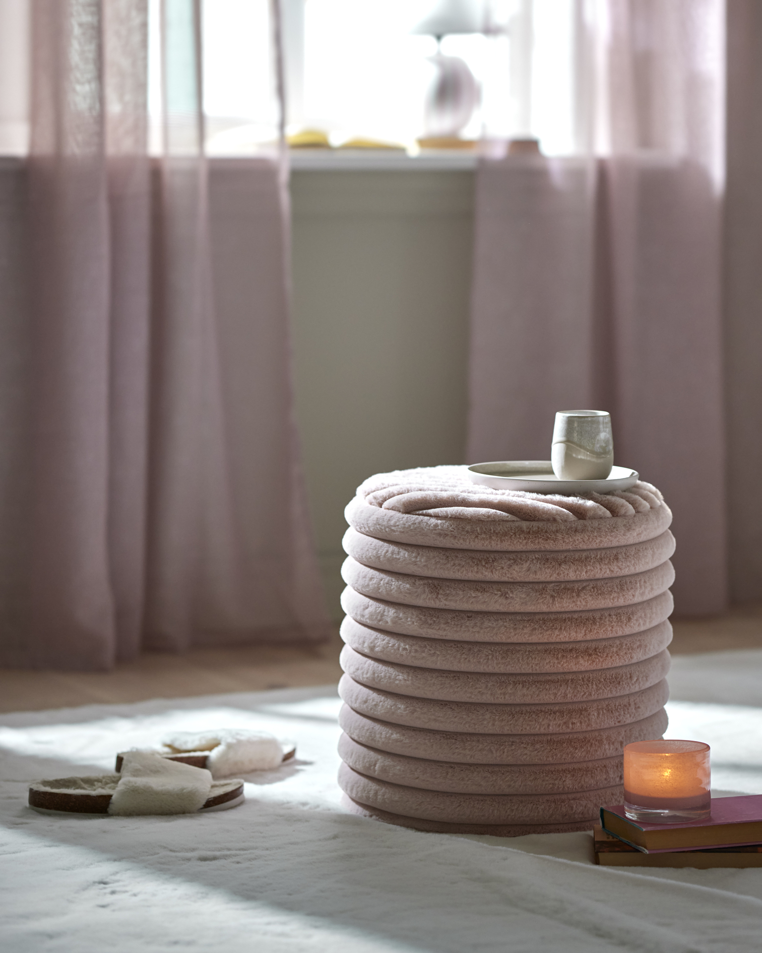 Rosa puff av mjuk konstpäls står på en vit matta tillsammans med ett par tofflor och en ljuslykta på några böcker.