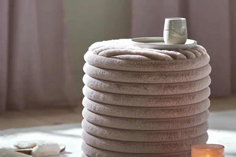 Rosa puff av mjuk konstpäls står på en vit matta tillsammans med ett par tofflor och en ljuslykta på några böcker.