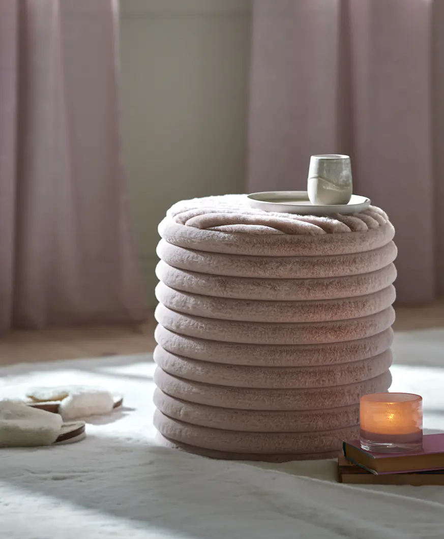 Rosa puff av mjuk konstpäls står på en vit matta tillsammans med ett par tofflor och en ljuslykta på några böcker.