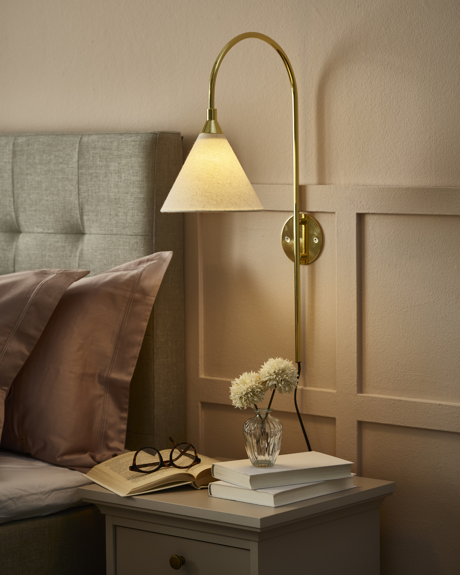 En guldig lampa med vit skärm sitter på väggen bredvid en säng.