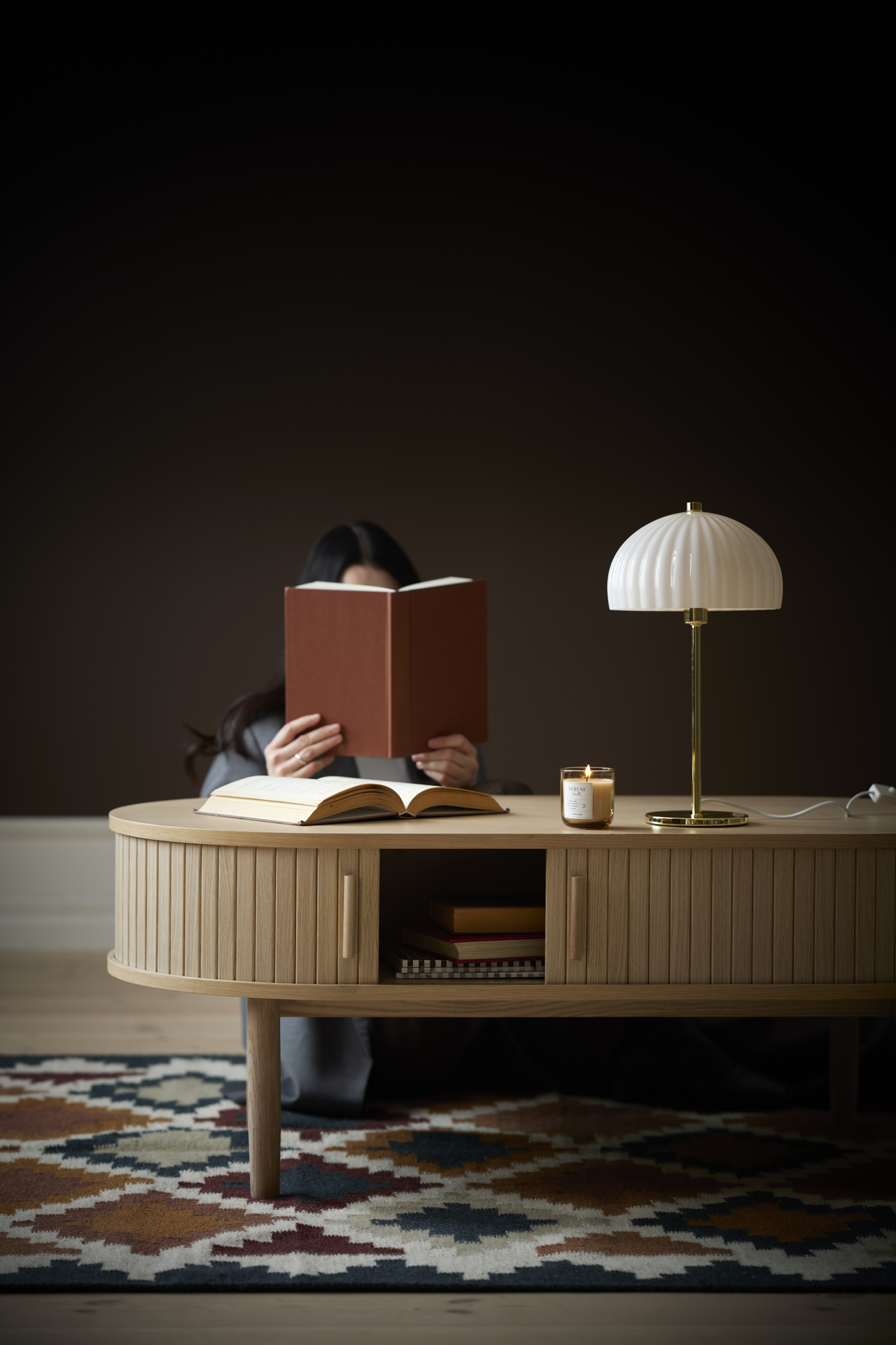 En kvinna sitter bakom ett soffbord och läser en bok. På bordet står en guldig lampa med vit skärm.