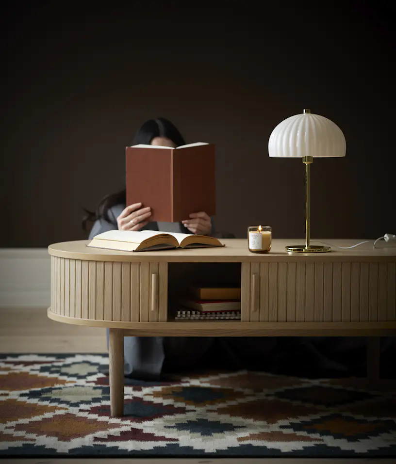 En kvinna sitter bakom ett soffbord och läser en bok. På bordet står en guldig lampa med vit skärm.