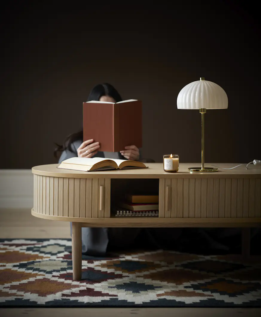 En kvinna sitter bakom ett soffbord och läser en bok. På bordet står en guldig lampa med vit skärm.
