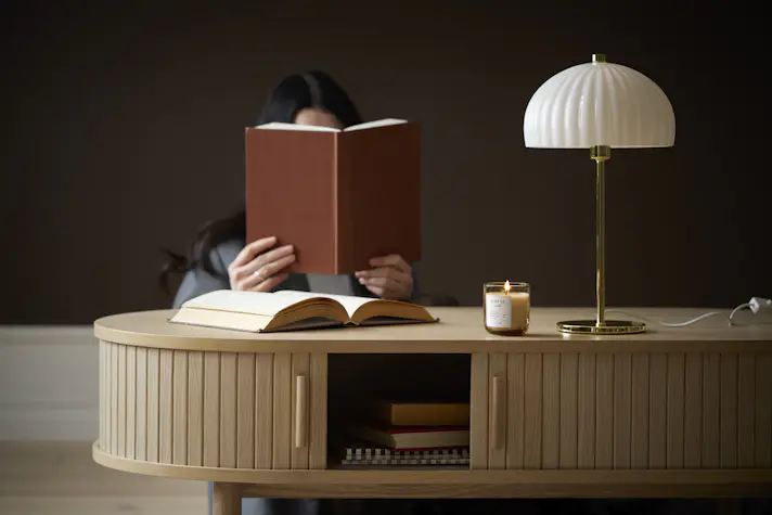 En kvinna sitter bakom ett soffbord och läser en bok. På bordet står en guldig lampa med vit skärm.