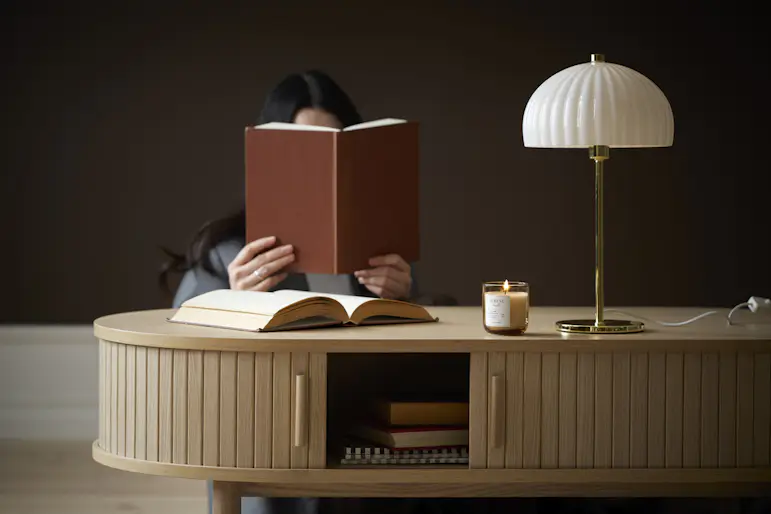 En kvinna sitter bakom ett soffbord och läser en bok. På bordet står en guldig lampa med vit skärm.
