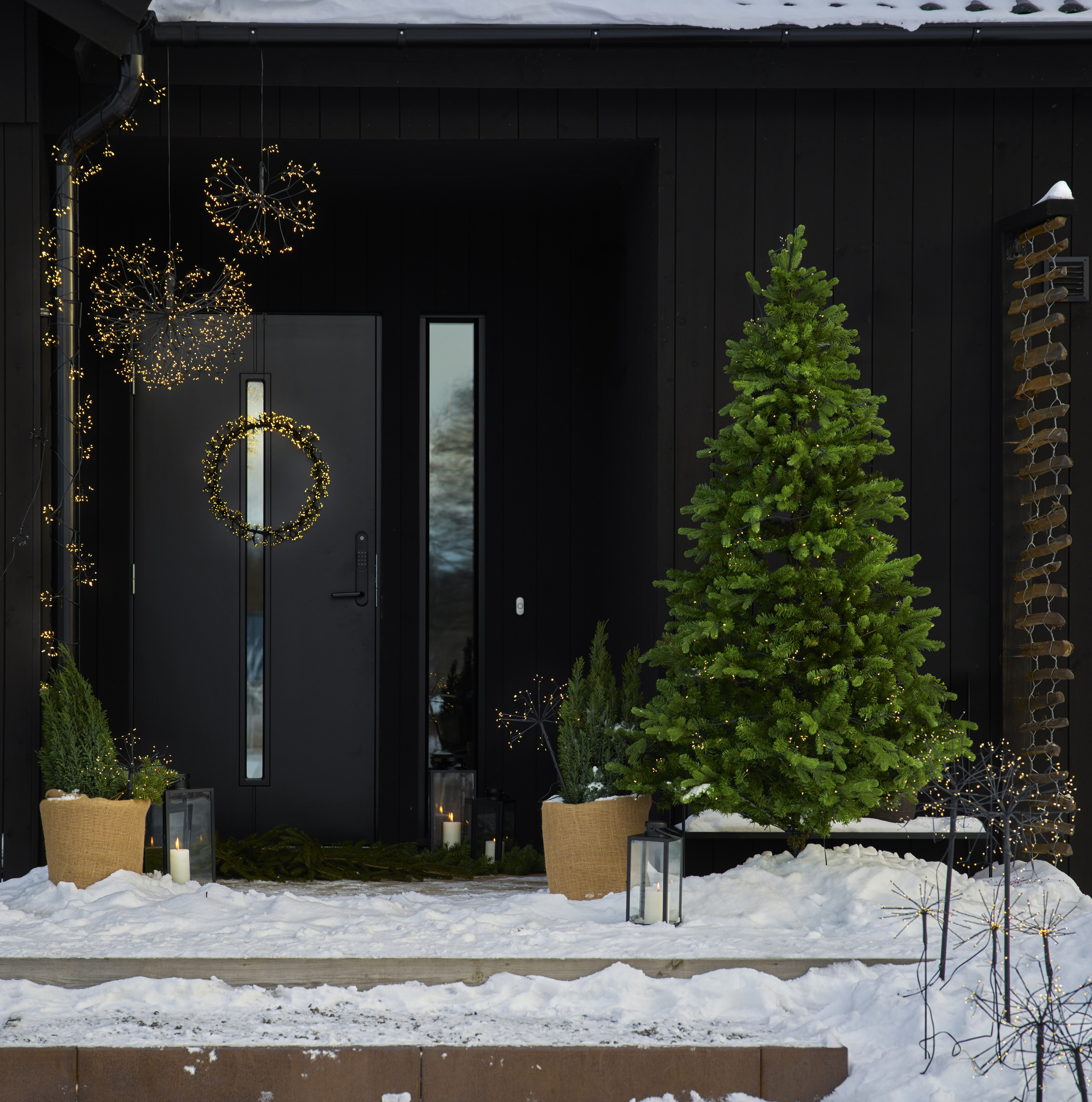 En snötäckt entré är dekorerad med ljusslingor, ljusbollar, en ljuskrans och en julgran med belysning.