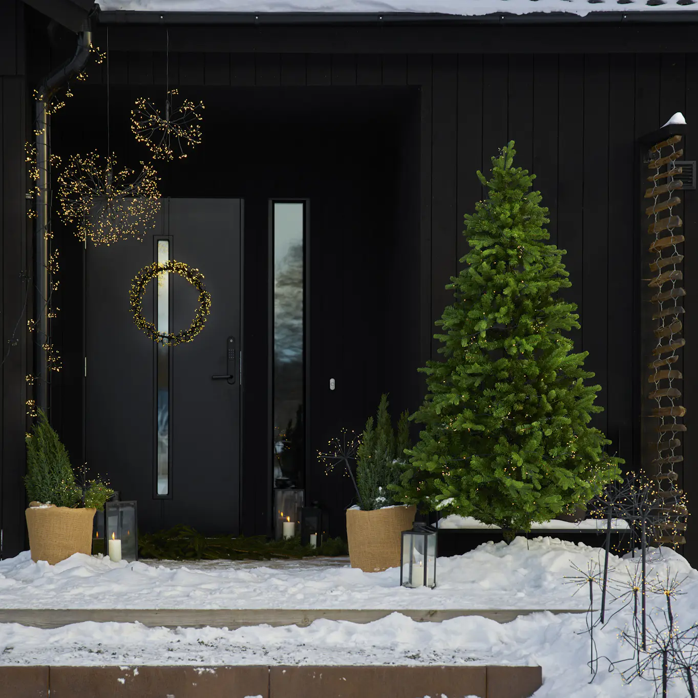 En snötäckt entré är dekorerad med ljusslingor, ljusbollar, en ljuskrans och en julgran med belysning.