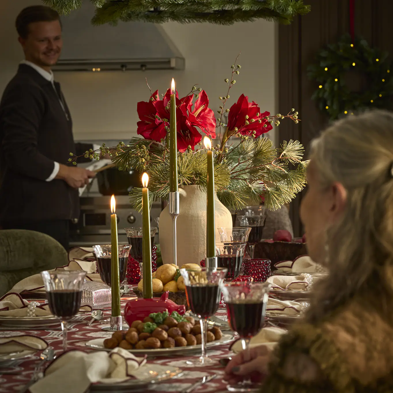 Ett juldukat matbord i röda och gröna toner. I förgrunden syns en kvinna hålla i ett vinglas och i bakgrunden står en man vid spisen.
