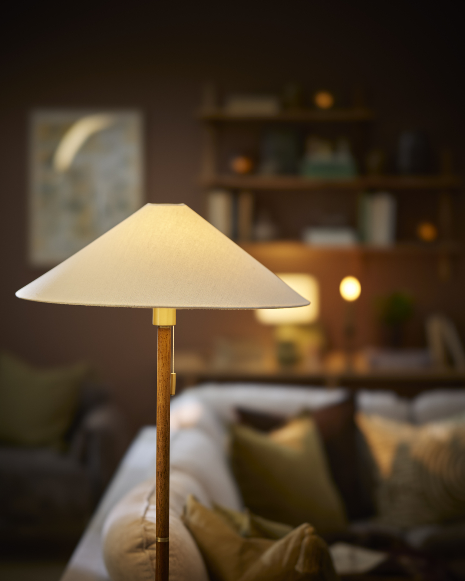 En golvlampa av trä med en vit lampskärm står i en mörk vardagsrumsmiljö.