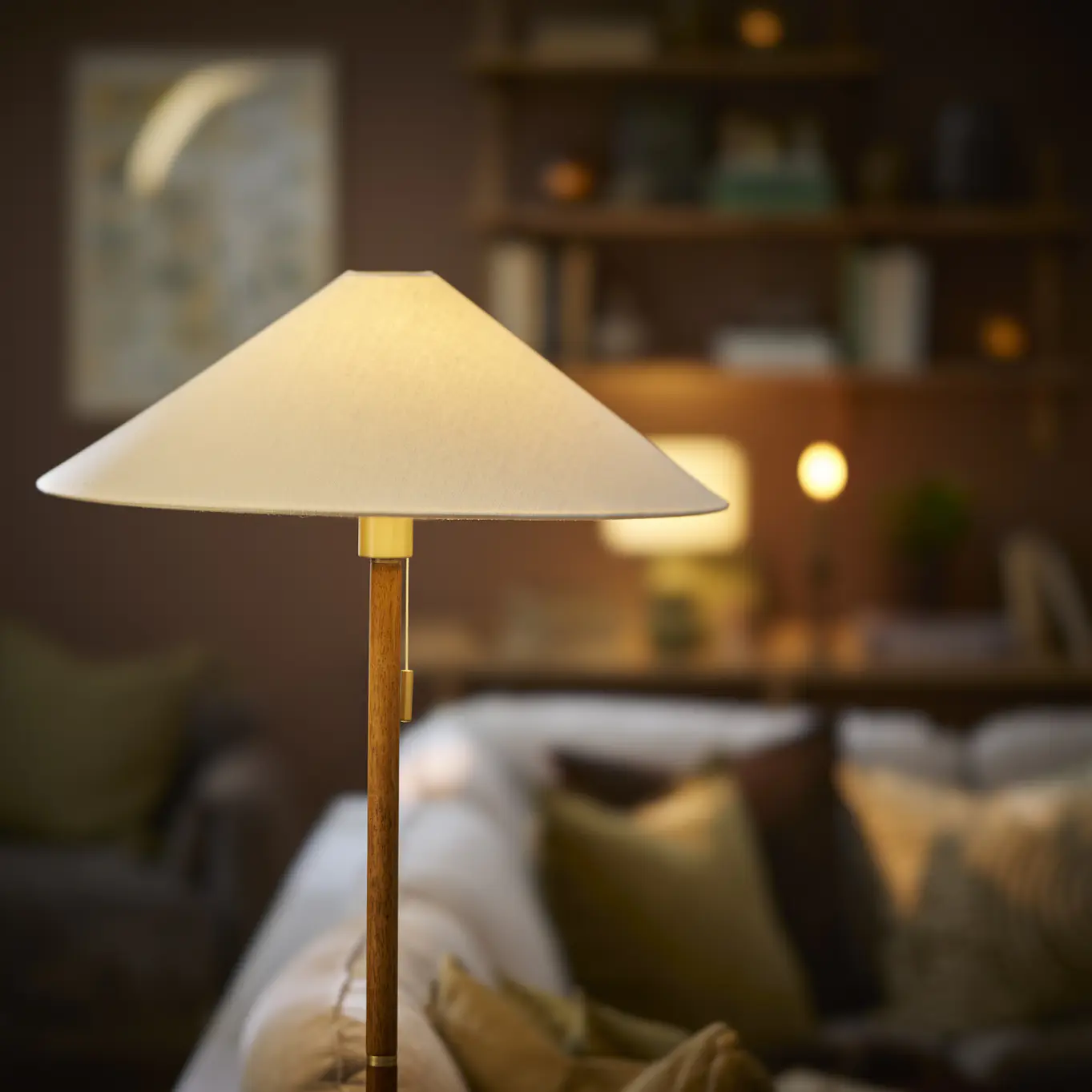 En golvlampa av trä med en vit lampskärm står i en mörk vardagsrumsmiljö.