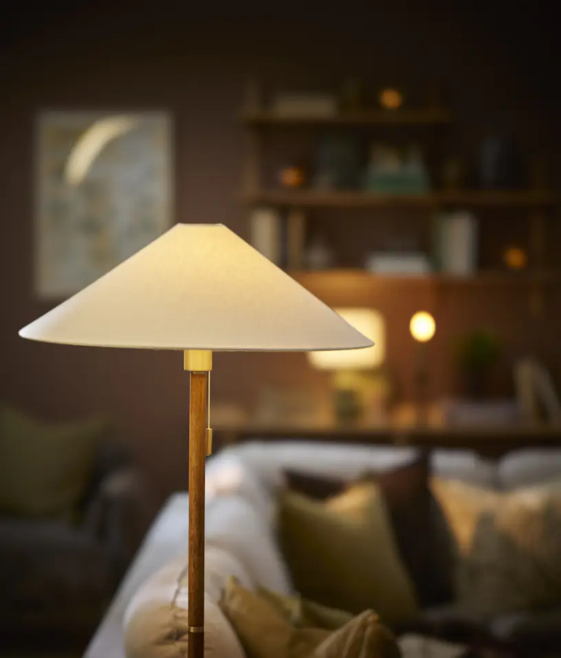 En golvlampa av trä med en vit lampskärm står i en mörk vardagsrumsmiljö.