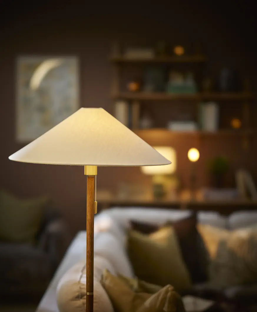 En golvlampa av trä med en vit lampskärm står i en mörk vardagsrumsmiljö.