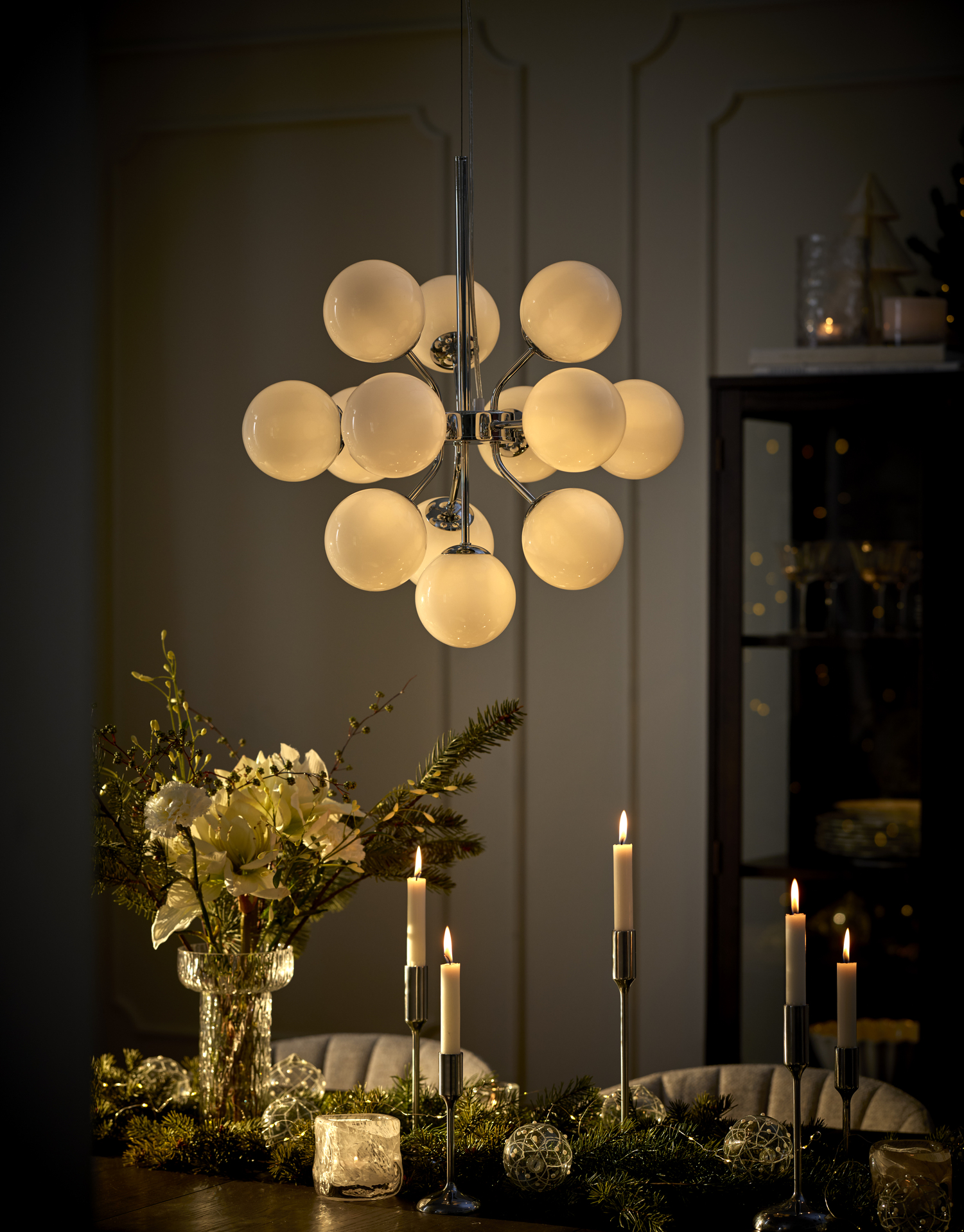 En taklama i krom och med flera glaskulor hänger över ett bord där det står en vas med amaryllis och flera ljusstakar med levande ljus.