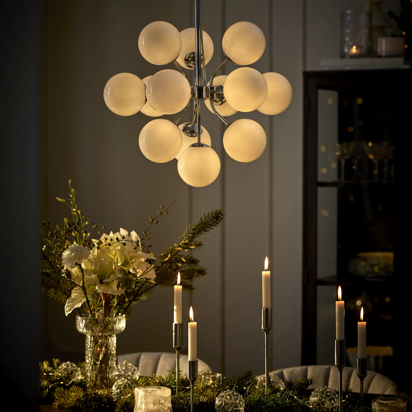 En taklama i krom och med flera glaskulor hänger över ett bord där det står en vas med amaryllis och flera ljusstakar med levande ljus.